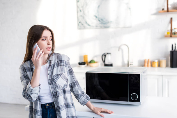shocked woman talking on smartphone near microwave in kitchen  - Photo, Image