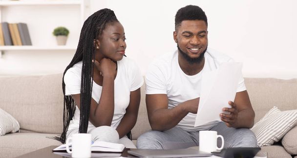 Smiling black couple reading insurance papers at home, checking agreement details - 写真・画像