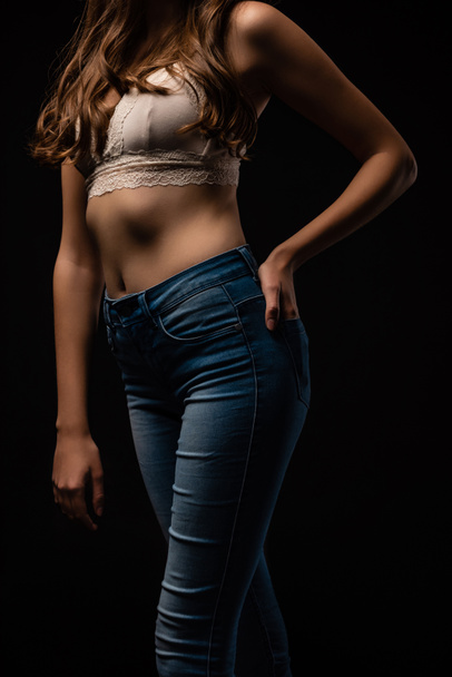 abgeschnittene Ansicht von sexy jungen Mädchen in BH und Jeans posiert mit der Hand in der Tasche isoliert auf schwarz - Foto, Bild