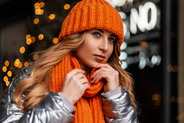 Leuke jonge vrouw in een oranje hoed met een warme gebreide sjaal in een jeugdige glanzende jas poseert in de stad en droomt van een nieuwjaarswonder. Portret van een mooi meisje bij een etalage met lichtjes. - Foto, afbeelding