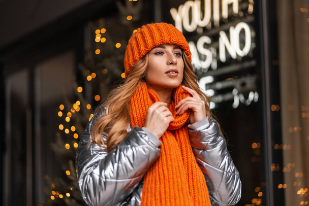 Ładna miejska kobieta z fryzurą w szydełkowym, stylowym kapeluszu z pomarańczowym ciepłym szalikiem w modnej, błyszczącej kurtce, pozującej w mieście, w pobliżu nowoczesnego sklepu. Europejska dziewczyna w modnej zimowej sukience. - Zdjęcie, obraz