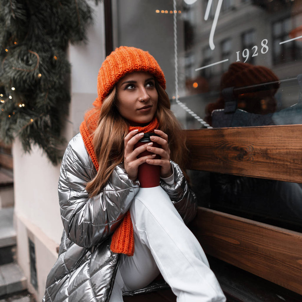 Boldog aranyos fiatal nő egy kötött téli elegáns kalap egy narancssárga sál egy csillogó fényes kabát ül az ablak mellett, és az álmok. Gyönyörű vörös hajú lány élvezi a hétvégét az utcán. - Fotó, kép