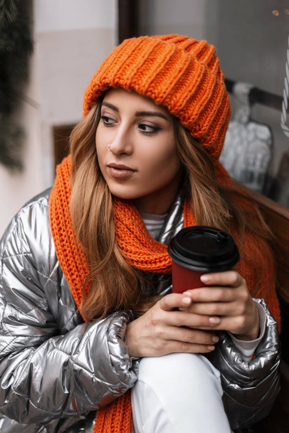 Portrait d'une jeune femme au chapeau orange à la mode avec une écharpe dans une veste glamour brillante d'hiver près d'une fenêtre vintage. Belle fille modèle s'assied avec une tasse de thé délicieux et profite d'un repos
. - Photo, image