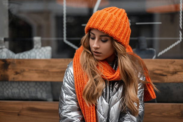 Pretty beautiful young woman with red hair in a brilliant trendy jacket in a knitted orange hat with a stylish scarf sits and looks down near a window in the city. Attractive fashionable girl model. - Photo, Image