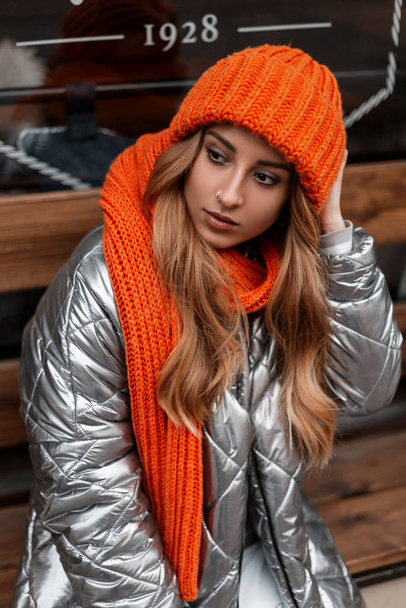 Ευρωπαϊκή κοκκινομάλλα νεαρή γυναίκα σε ένα μοντέρνο πορτοκαλί πλεκτό καπέλο σε ένα κομψό χειμωνιάτικο μπουφάν που ποζάρει κοντά σε έναν γυάλινο τοίχο στην πόλη. Χαριτωμένο κορίτσι με μοντέρνα ρούχα που ποζάρει στο δρόμο. Απλή εμφάνιση. - Φωτογραφία, εικόνα