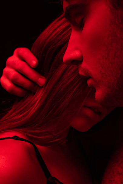 sexy jeune homme avec les yeux fermés touchant fille cheveux en lumière rouge isolé sur noir
 - Photo, image