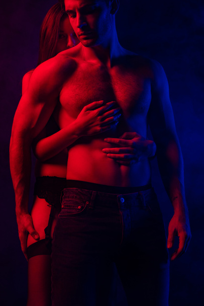 passionate undressed sexy young couple embracing in red and blue light on black background - Photo, Image