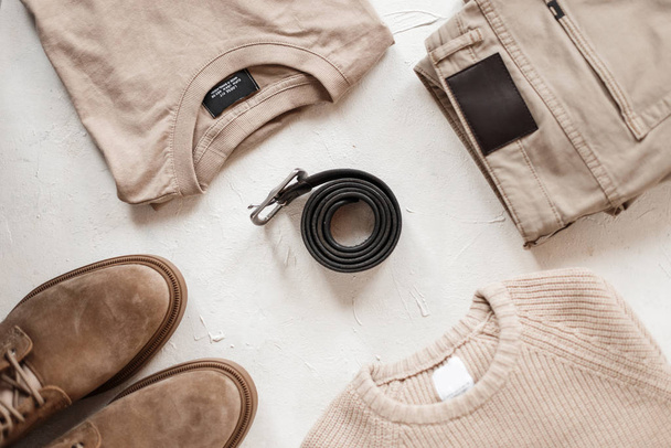 Top view on leather black men's stylish belt with vintage buckle on a white table among casual fashion clothes and shoes in beige colors. Trendy spring collection for men. Classic style. Close-up. - 写真・画像