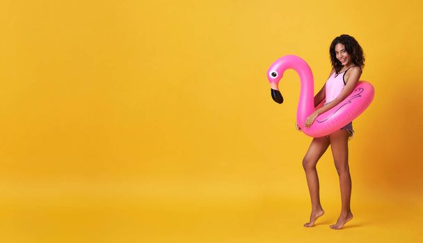 Happy smiling african woman dressed in swimwear holding flamingo rubber ring and enjoying their summer vacation getaway in yellow banner background with copy space. - Foto, Imagem