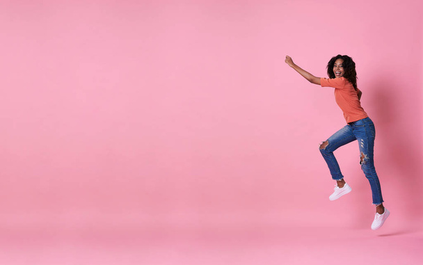Joven mujer africana alegre en camisa naranja saltando y celebrando sobre fondo de bandera rosa con espacio de copia
. - Foto, Imagen
