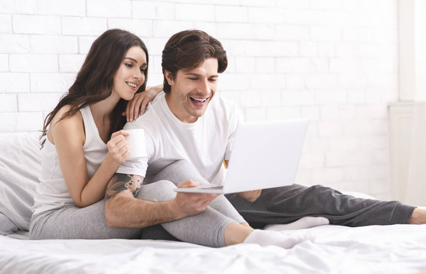 興奮した配偶者はノートパソコンで情報を楽しんで、ベッドで休んでいます - 写真・画像