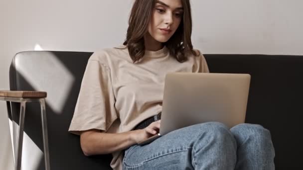 Smiling brunette woman using laptop computer while sitting on sofa indoors - Filmmaterial, Video
