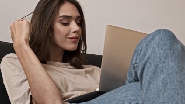 Άποψη από τα κάτω της Happy μελαχρινή γυναίκα σκέφτεται κάτι και παίζει με τα μαλλιά της, ενώ κάθεται με φορητό υπολογιστή σε εσωτερικό καναπέ - Πλάνα, βίντεο