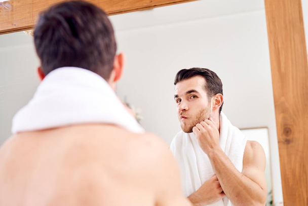 Unshaven male with white towel on his neck standing near mirror - Foto, afbeelding