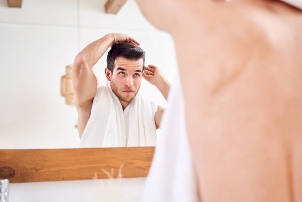 Young man combing his hair while standing with white towel on neck near mirror in bathroom - Fotó, kép