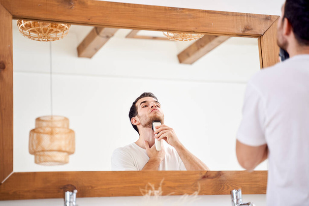 Brunet man in white T-shirt shaves while standing near mirror in bath - Foto, Bild