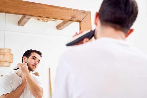 Close up brunet male in white T-shirt shaves while standing near mirror in bath - Foto, Bild