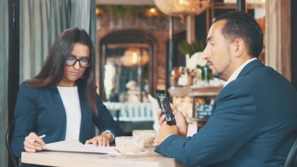Two businesspeople work in a cafe. Woman is learning attentively the documents. Man is on video chat. Side view. Close up. Copy space. - Imágenes, Vídeo