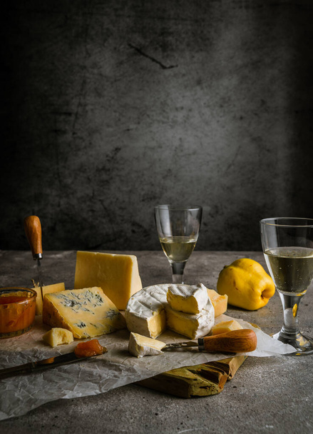 Low key still life with various cheeses, wine in glasses, quince fruit, and jam, with copy space - Photo, Image