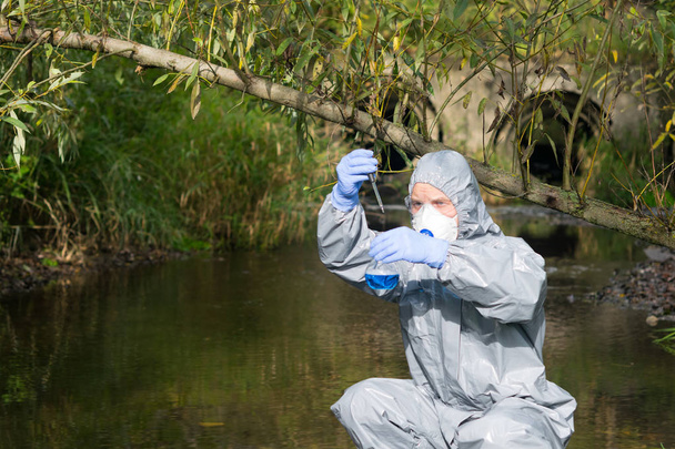 a scientist in a protective suit and mask collects a sample of water in a flask with a blue liquid - Foto, Imagem