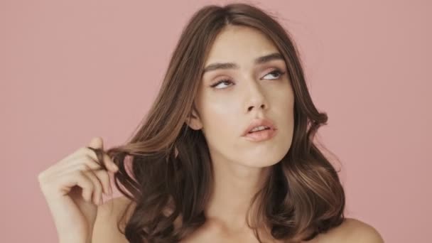 Beautiful pensive half-naked woman is twisting a curl of hair on a finger while thinking about something isolated over pink background - Кадры, видео