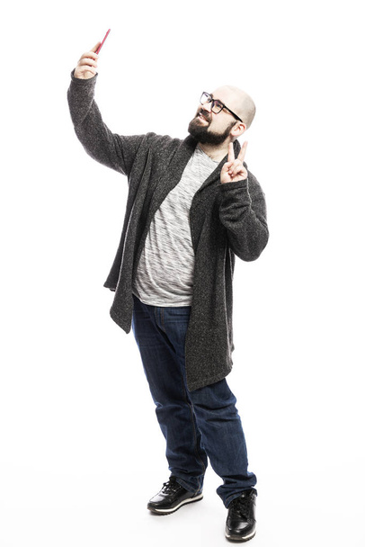 A bald man with glasses and a beard takes a selfie on the phone. Full height. Isolated on a white background.  - Valokuva, kuva