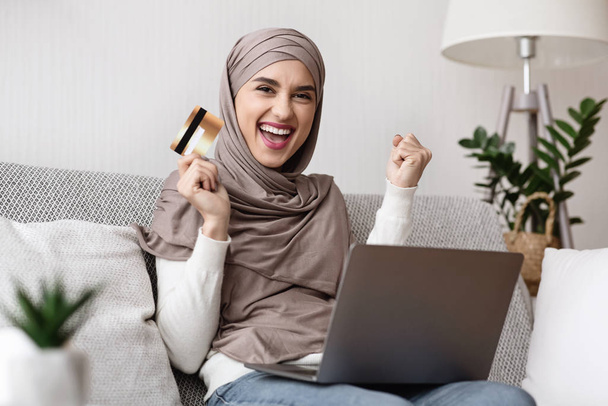 Chica musulmana alegre celebrando el éxito con tarjeta de crédito y portátil
 - Foto, Imagen