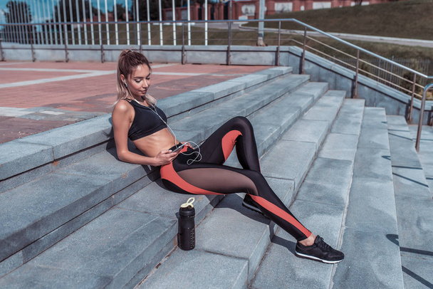 woman resting after workout, summer city, sportswear, mobile phone headphones, listening music in application. Bottle shaker with protein. Reads a message, sending SMS on social networks. - Zdjęcie, obraz