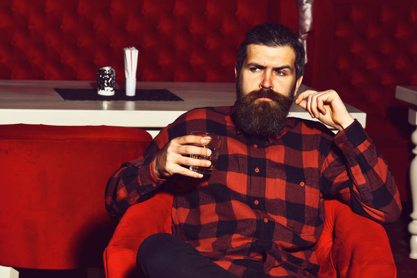 bearded man hipster holding glass of whiskey in red chair - Photo, Image