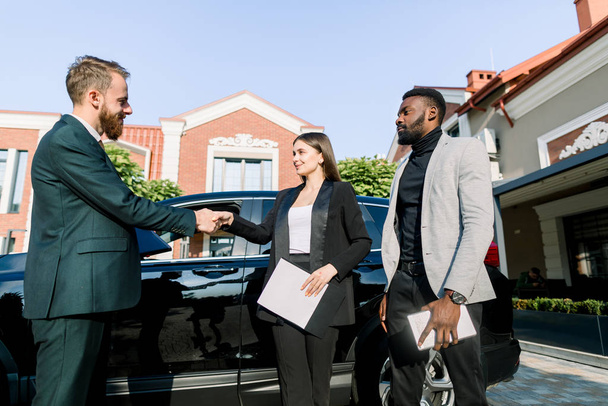 Welcome to our team. Three young multiethnical business people standing near car outdoors. Happy young woman shaking hands with new colleague man, African man holding tablet stands nearby - Foto, Imagen