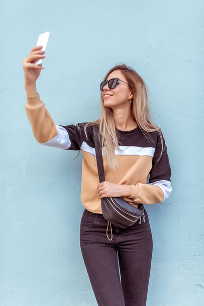 Beautiful girl summer city, hand mobile phone, beige sweater black glasses. Photographs smartphone, selfie phone, happy smiling laughs. Post social networks. Modern fashion style woman. - Foto, afbeelding