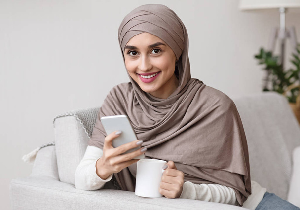 Piękna arabska dziewczyna w hidżabie ze smartfonem i filiżanką herbaty - Zdjęcie, obraz