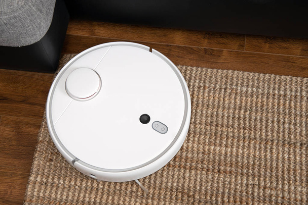 Ρομποτική ηλεκτρική σκούπα τρέχει κάτω από καναπέ στο δωμάτιο σε laminate πάτωμα σύγχρονη έξυπνη τεχνολογία καθαρισμού housekeping. - Φωτογραφία, εικόνα