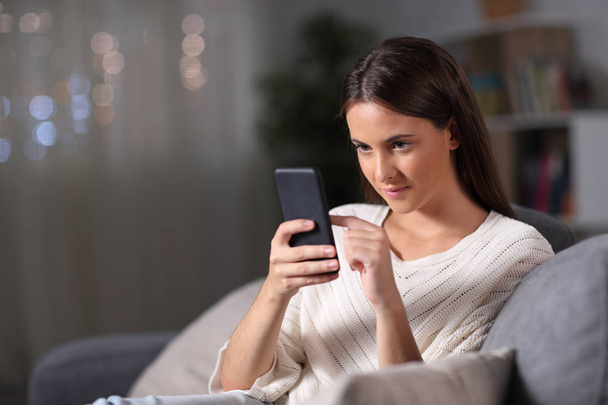 Девушка проверяет содержание мобильного телефона, сидя ночью дома
 - Фото, изображение