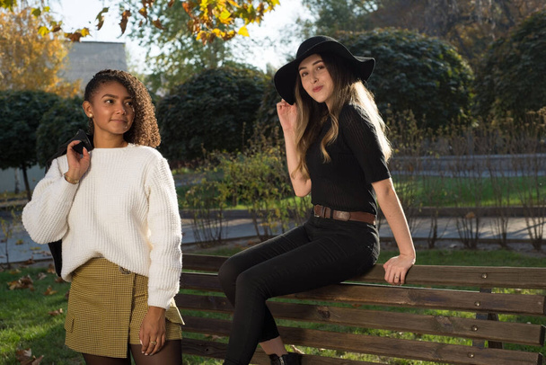 pair of young beautiful mexican generation y girls. The girl is sitting on the back of the bench, a girlfriend is standing next to curly hair and a white sweater, holding a jacket on her shoulder - 写真・画像