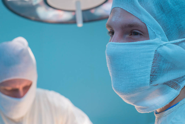 手術室の外科医の下のビューでは、滅菌マスクで、外科的介入の準備をし、腕を上げ、滅菌手袋で。灯の背景には - 写真・画像