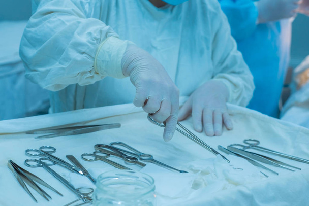 közelkép, egy sebészeti nővér kezei, amint orvosi műszereket vesz ki egy steril asztalról, egy műtétet végző orvos csapat hátterében. - Fotó, kép