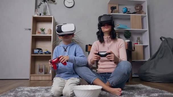 Madre e hijo modernos de buen aspecto con gafas 3D virtuales especiales sentados en la alfombra y jugando a videojuegos
 - Imágenes, Vídeo