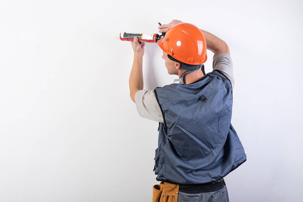 Builder with sealant. In work clothes and hard hat. On a light gray background. - Photo, Image