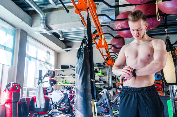 A man in a gym without a T-shirt stands in front of a window and looks at his watch - Valokuva, kuva