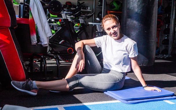 Ein Mädchen in weißem T-Shirt und grauer Leggings sitzt in einem Fitnessclub auf einer Gymnastikmatte, lächelt und blickt in die Kamera.  - Foto, Bild