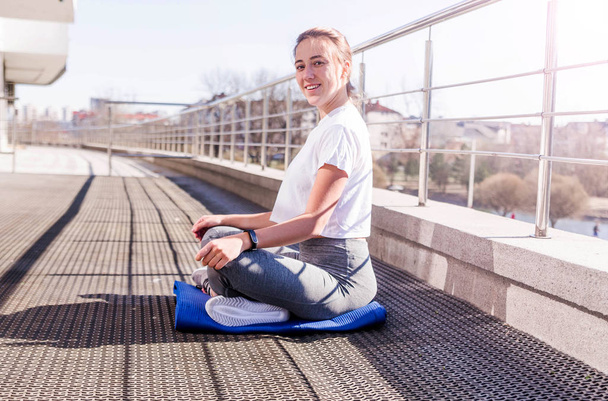 Young athletic woman in a white T-shirt and gray leggings sits in a lotus position in the fresh air in sunny weather on the terrace. Smile and look at camera - Fotografie, Obrázek