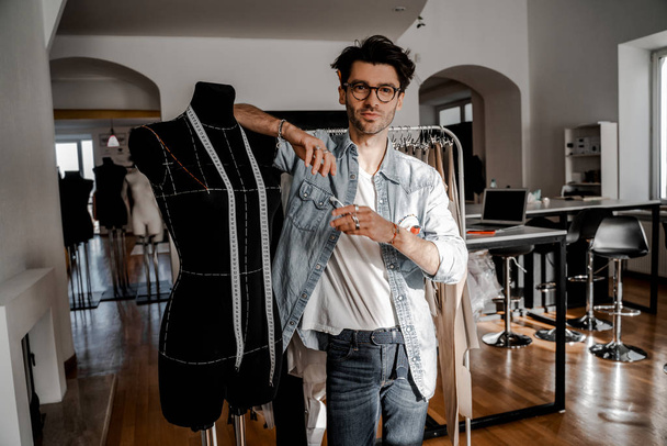 Jóképű profi férfi elegáns ruhákat a design stúdió áll magabiztos és sikeres közelében egy próbababa. Szabó egy professzionális eszközzel a kezében. Fiatal és kreatív tervező - Fotó, kép