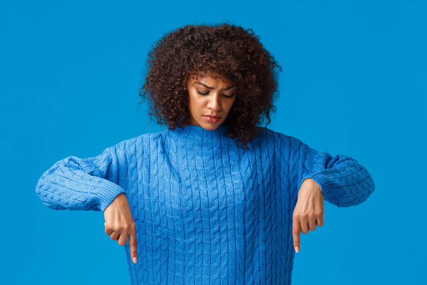 Serious-looking upset and gloomy african-american woman with afro haircut having sad winter holidays, looking and pointing fingers down, staring disappointed, standing blue background - Fotó, kép