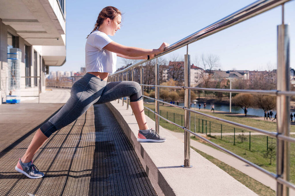 A cheerful girl is suitable for a morning run. Young woman in a white T-shirt and gray leggings, leaning on a railing and stretching legs and ankles.  - Photo, Image