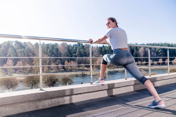 A cheerful girl is suitable for a morning run. Young woman in a white T-shirt and gray leggings, leaning on a railing and stretching legs and ankles.  - Photo, Image