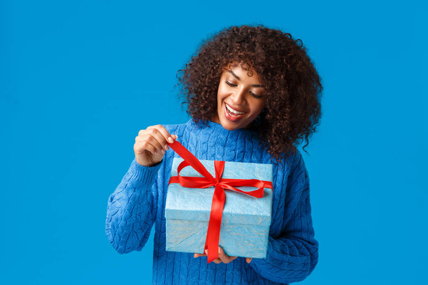 Meraklı ve mutlu, gülümseyen Afro-Amerikan kadın, kışın süveter giyen doğum günü kızı, hediyesini açmak için hediye düğümünü çekiyor ve içinde ne olduğunu görüyor, Noel 'i, yeni yılı, yeni yılı, mavi arka planı, - Fotoğraf, Görsel