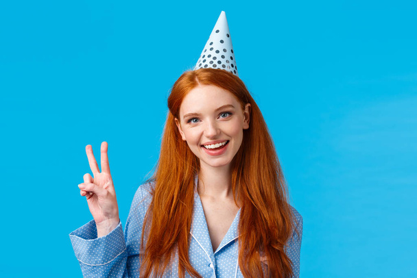 Lucky and enthusiastic feminine pretty redhead woman with long curly hair in nightwear, wearing birthday cap showing peace sign and smiling joyfully, celebrating b-day, blue background - Fotografie, Obrázek