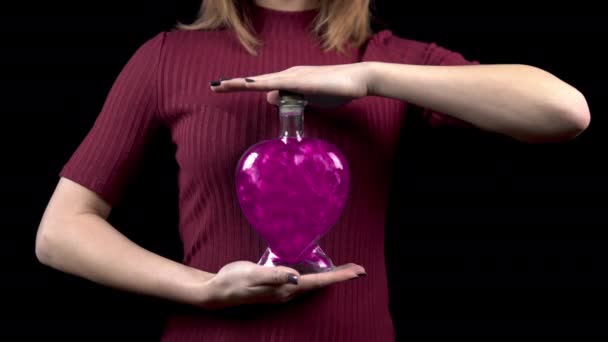 Een jonge vrouw houdt een fles met een liefdesdrank vast. Glazen fles in de vorm van een hart met roze vloeistof. Valentijnsdag is de dag van alle minnaars. - Video