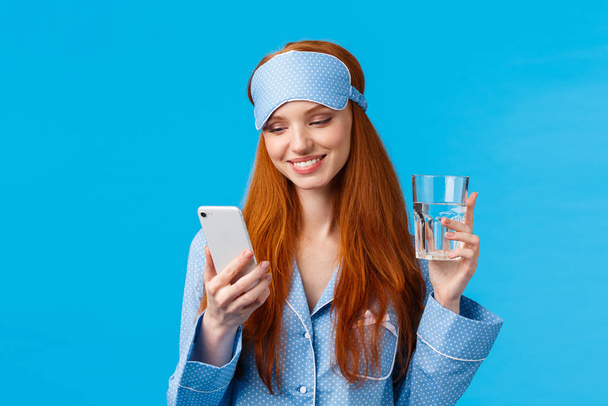 Happy and relaxed attractive feminine redhead woman holding smartphone and glass water, smiling joyfully and delighted as reading nice lovely message from mobile application, blue background - Photo, Image